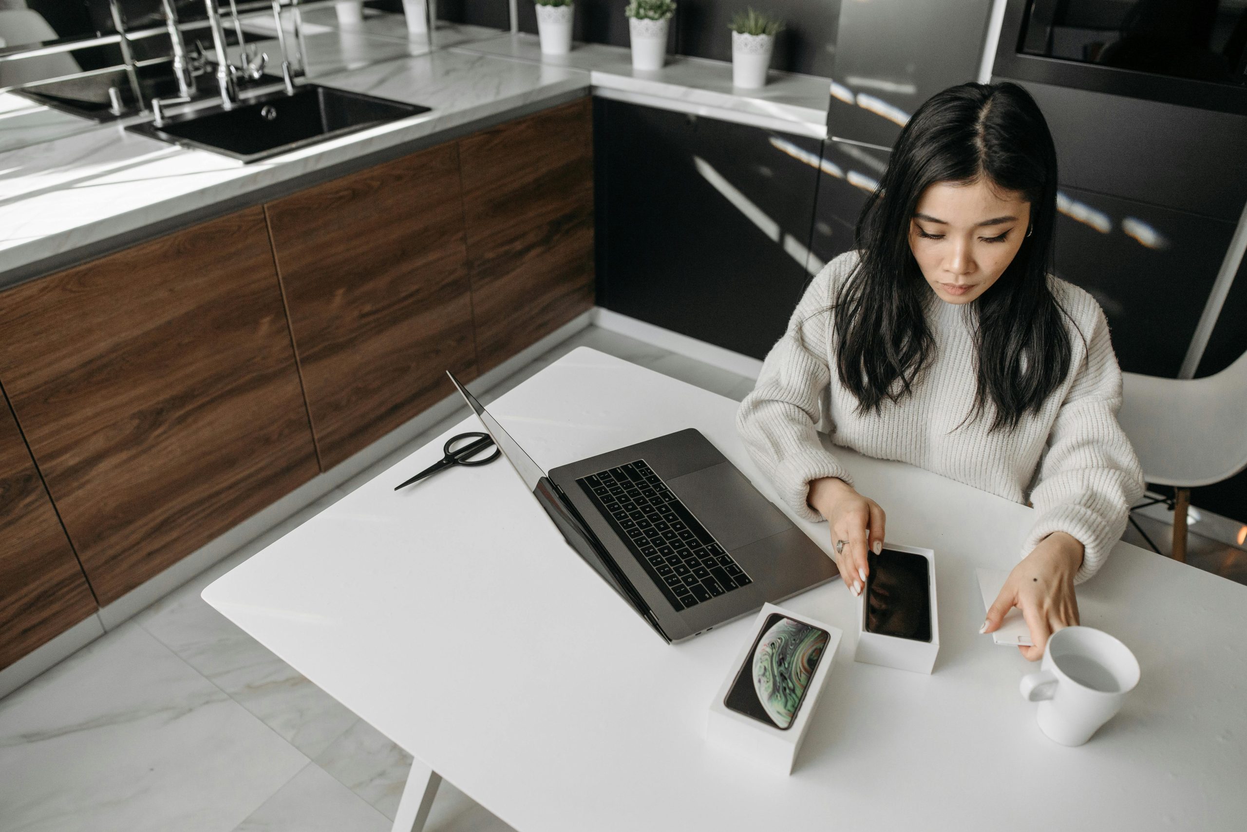 Woman unboxing a smartphone at a table with a laptop in a modern kitchen setting.